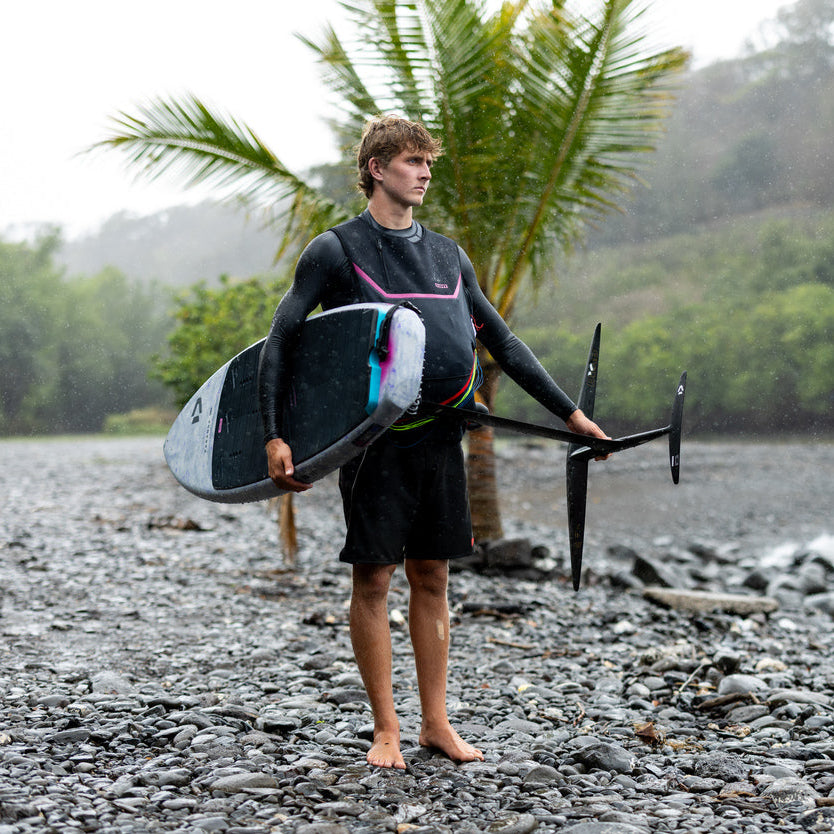 Person holding a Duotone Paradox Foil Board with a Duotone Crest Foil on a rocky beach with palm trees and water in the background
