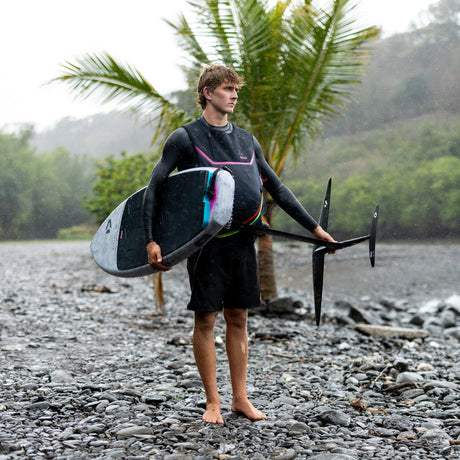 Person holding a Duotone Paradox Foil Board with a Duotone Crest Foil on a rocky beach with palm trees and water in the background