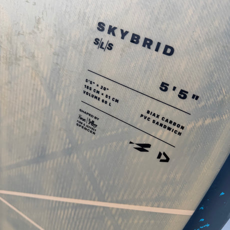 Close-up of a surfboard label reading SKYBRID S/L/S with dimensions 5'5" and material details, on a pale blue textured board surface.