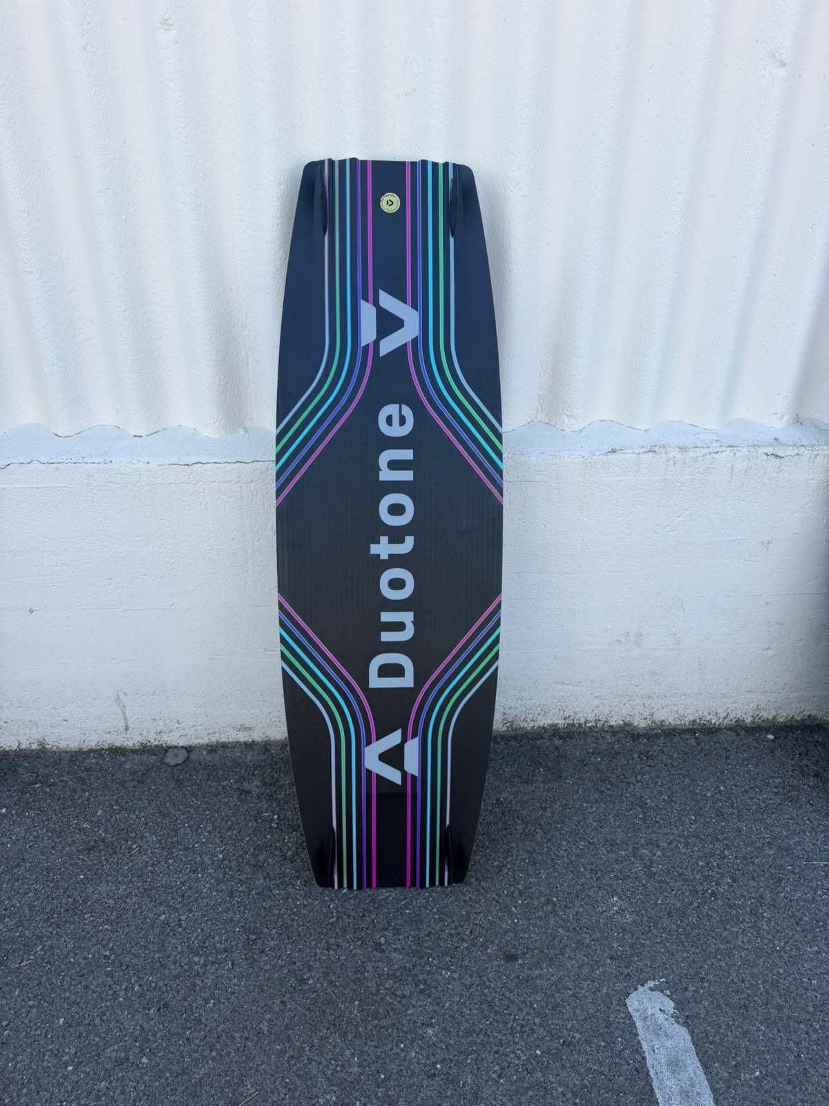 A Duotone wakeboard stands upright against a white corrugated metal wall on a gray asphalt surface. The board is black with vibrant multicolor neon stripes in pink, green, and blue, and features the Duotone brand name in white.