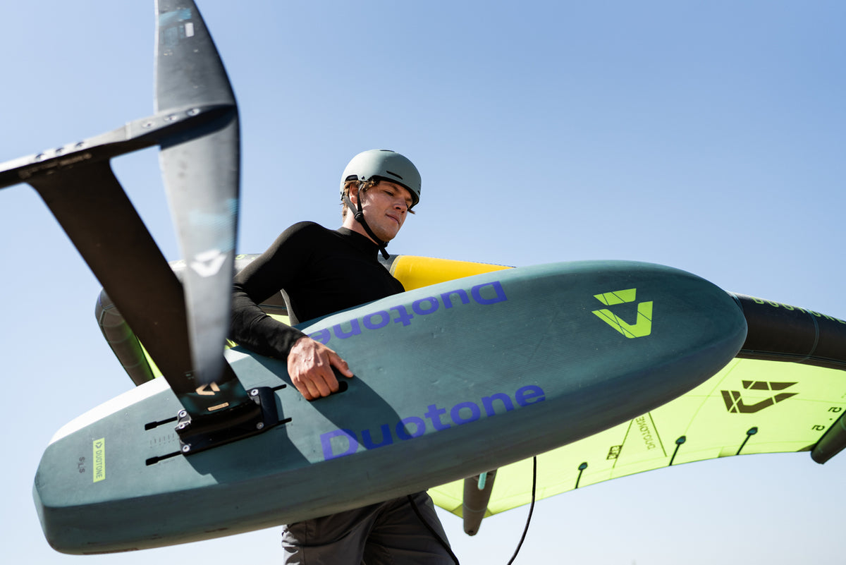 A person in a wetsuit and helmet carries a Duotone solar wing on a board against a clear blue sky.