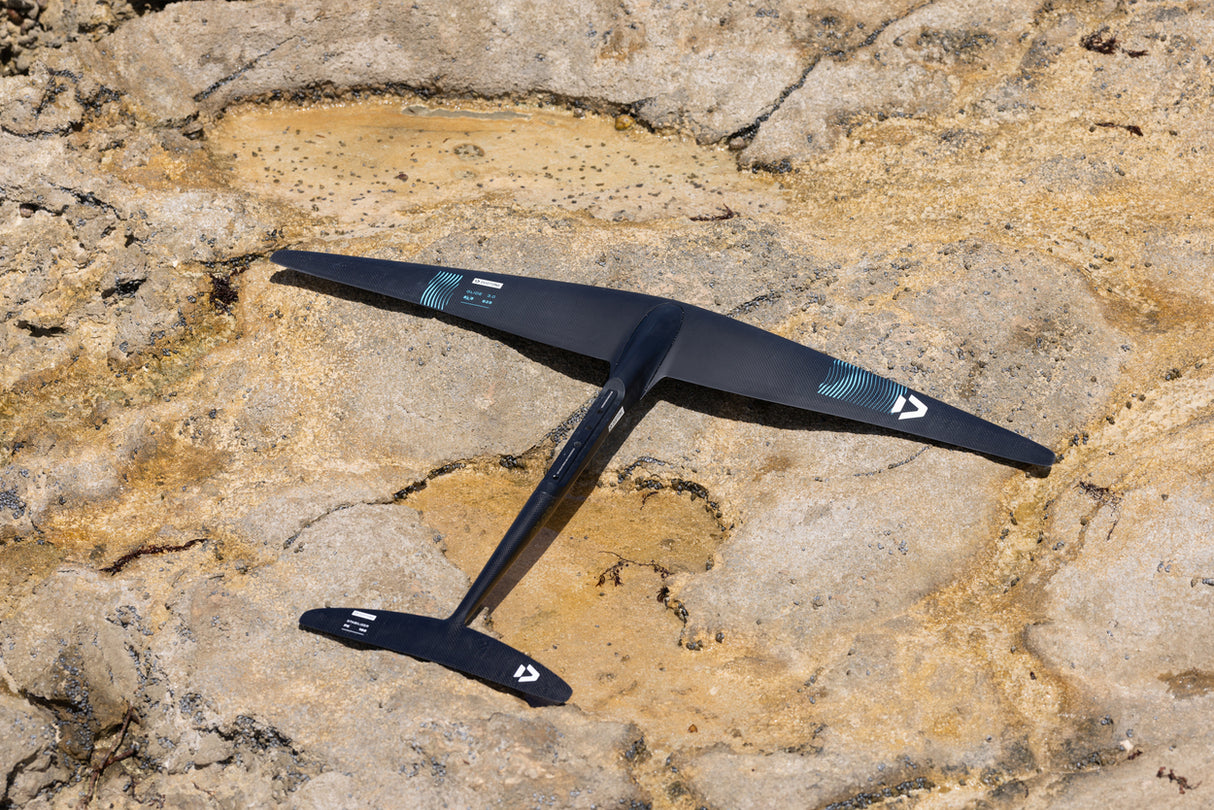 A black carbon-fiber ultralight glider with white logo markings rests diagonally on a textured, dry sandy rock surface.