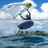 A windsurfer jumps high over blue ocean water under a bright sky with large white clouds. The surfer holds a large white sail with blue and yellow accents while riding a hydrofoil board.