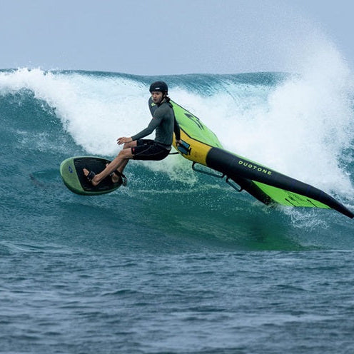 A surfer in a black wetsuit rides a duotone trace sls foil board over a breaking ocean wave, with white spray and blue water visible.
