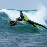 A surfer in a black wetsuit rides a duotone trace sls foil board over a breaking ocean wave, with white spray and blue water visible.