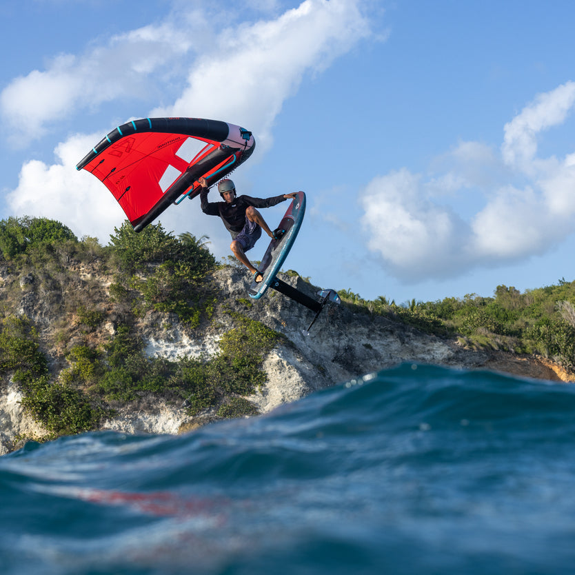 A wingsurfer jumps high over a rocky coastal cliff with blue ocean water in the foreground and a partly cloudy sky above. The wing is bright red with black and white sections, and the surfer wears dark clothing.